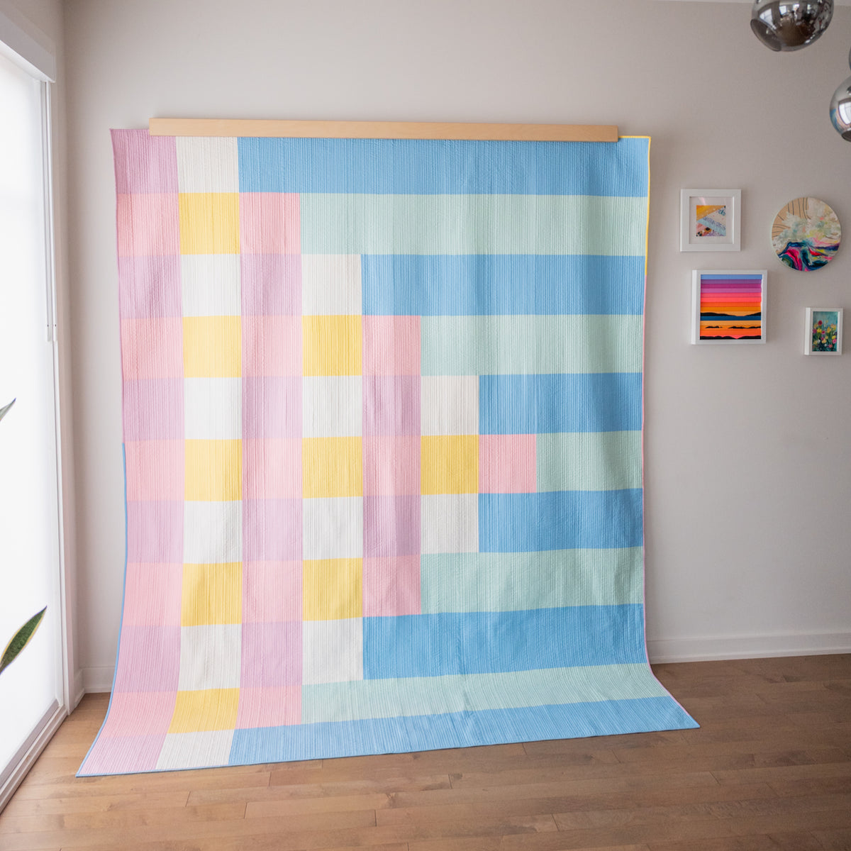 A large textile wall hanging or quilt with a pastel checkerboard block pattern, displayed on a wooden rod against a light gray wall in a room with wood flooring and small framed wall art.