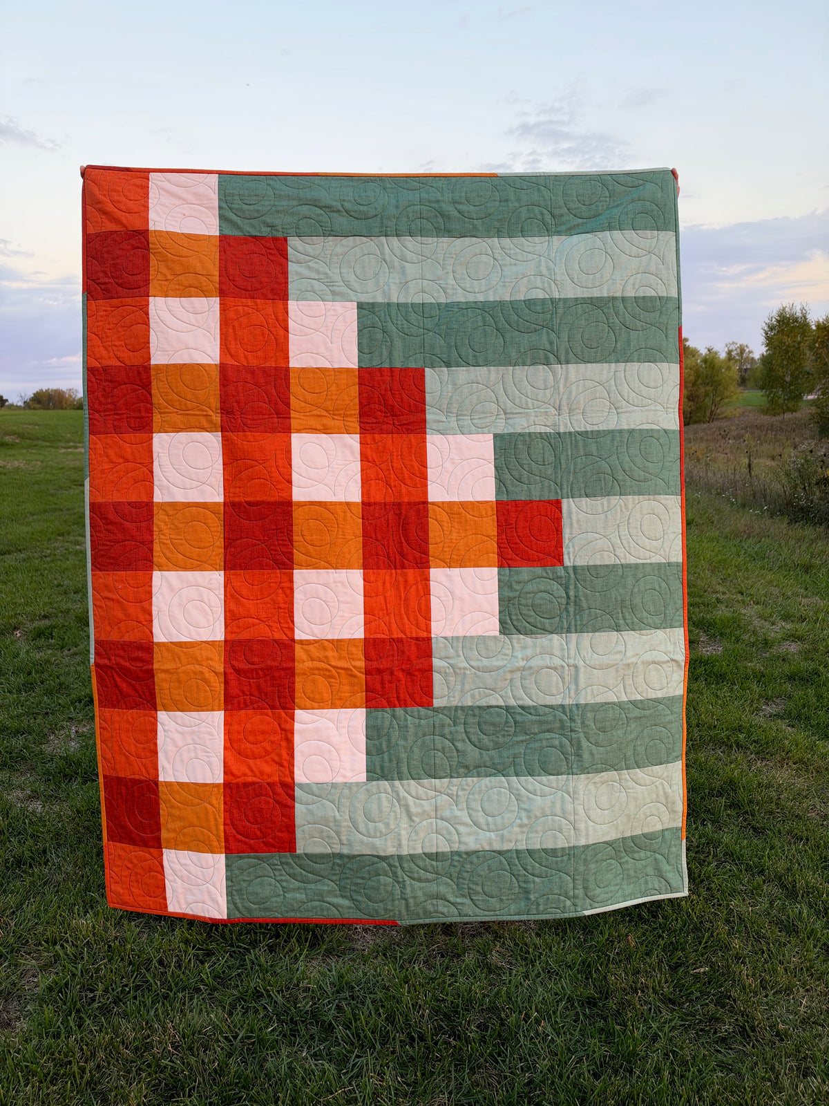 A handmade quilted patchwork blanket with a woven design, displayed outdoors on a grassy lawn during sunset.
